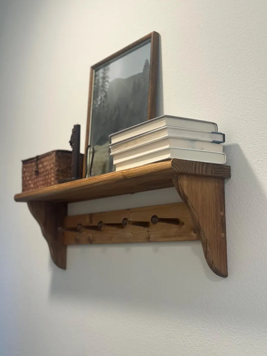 A Shaker peg shelf with stained pine ledge, curved brackets and wooden pegs, shown on a white wall with books and a framed print