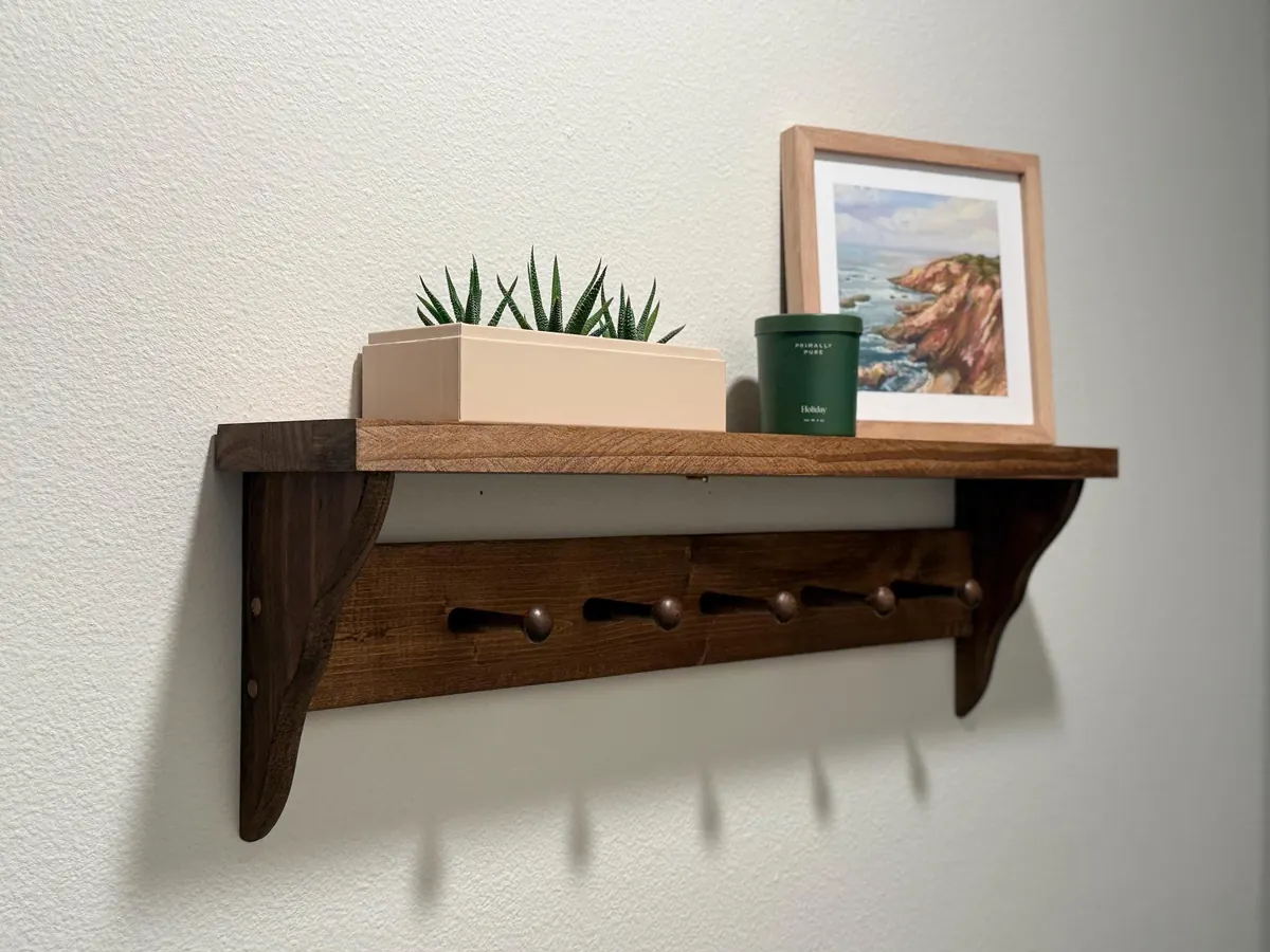A slim stained pine Shaker peg shelf on a wall with a small plant and a framed print on top