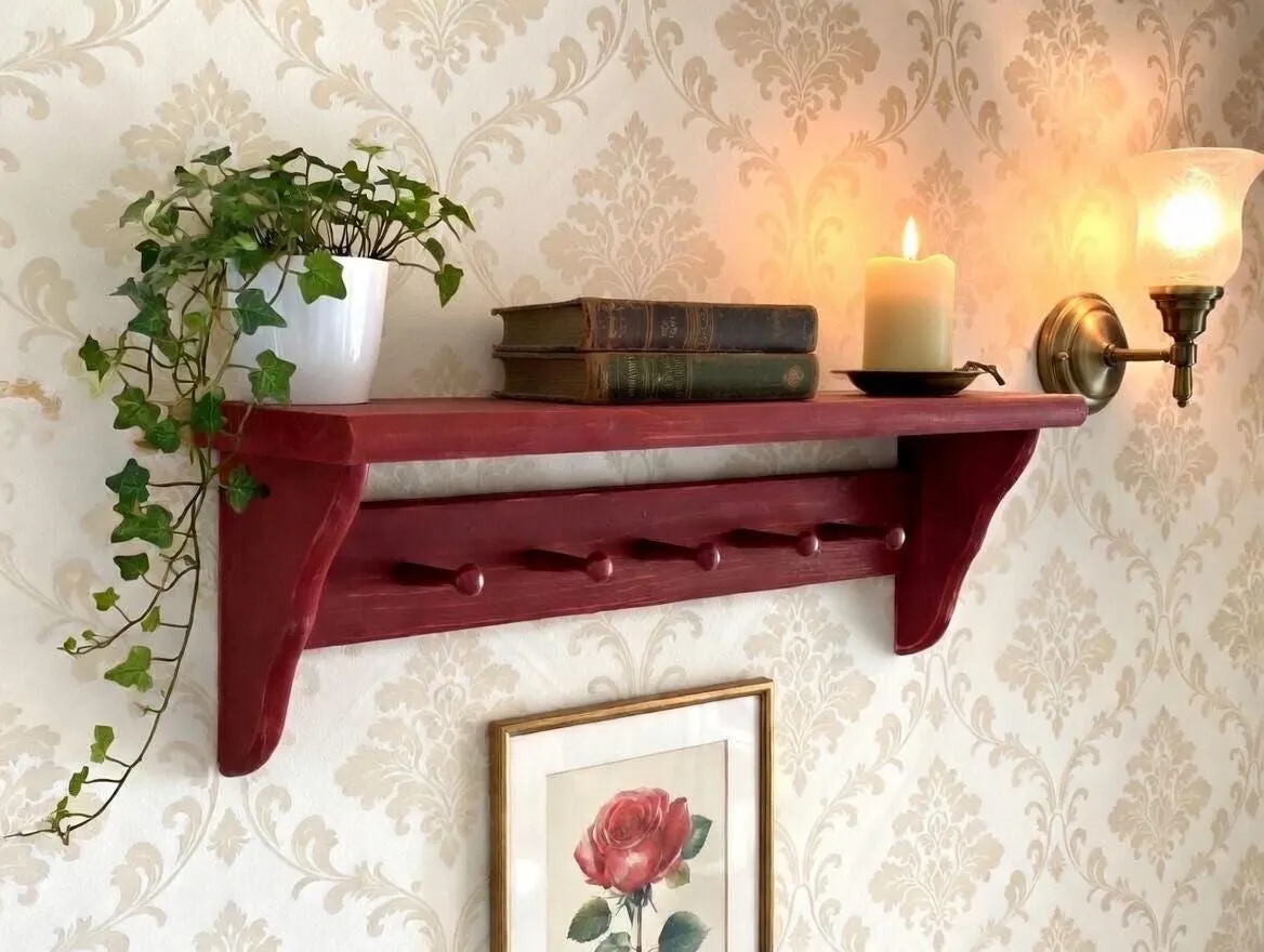 A distressed red Shaker peg shelf on floral wallpaper with an ivy plant, books and a candle on top