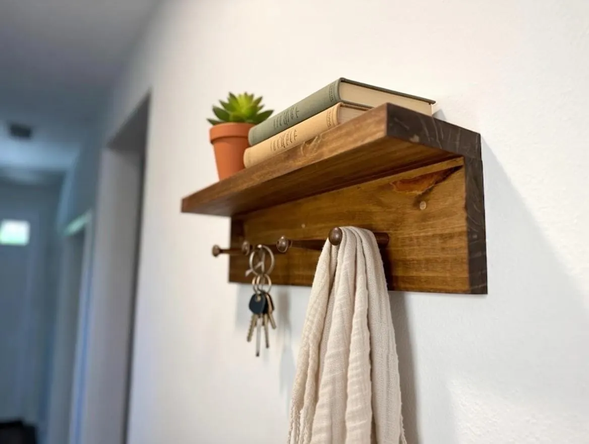 A rustic stained pine peg shelf in a hallway with keys and a linen towel hanging from the pegs and a small plant on top