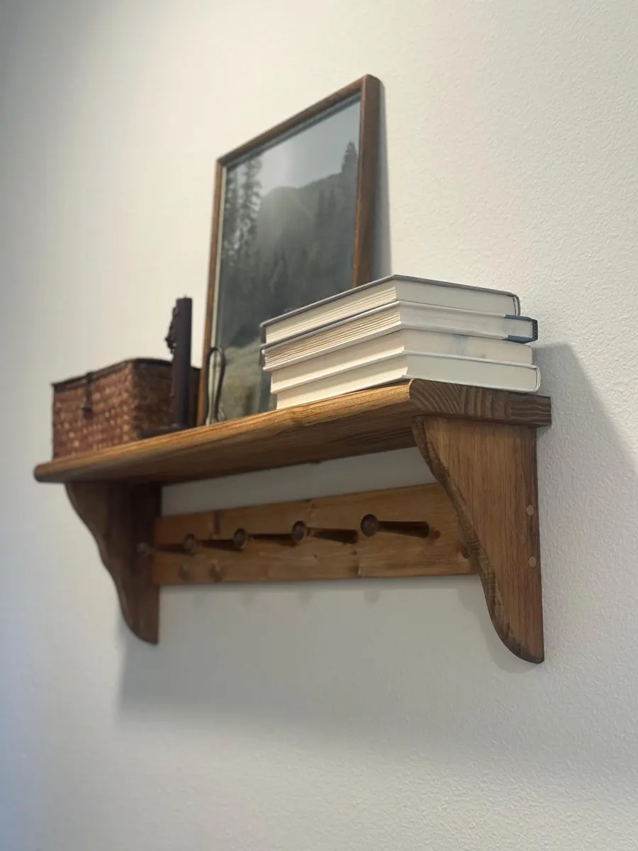 A stained pine peg shelf with a rounded-edge ledge holding a stack of books, a basket and a framed landscape print