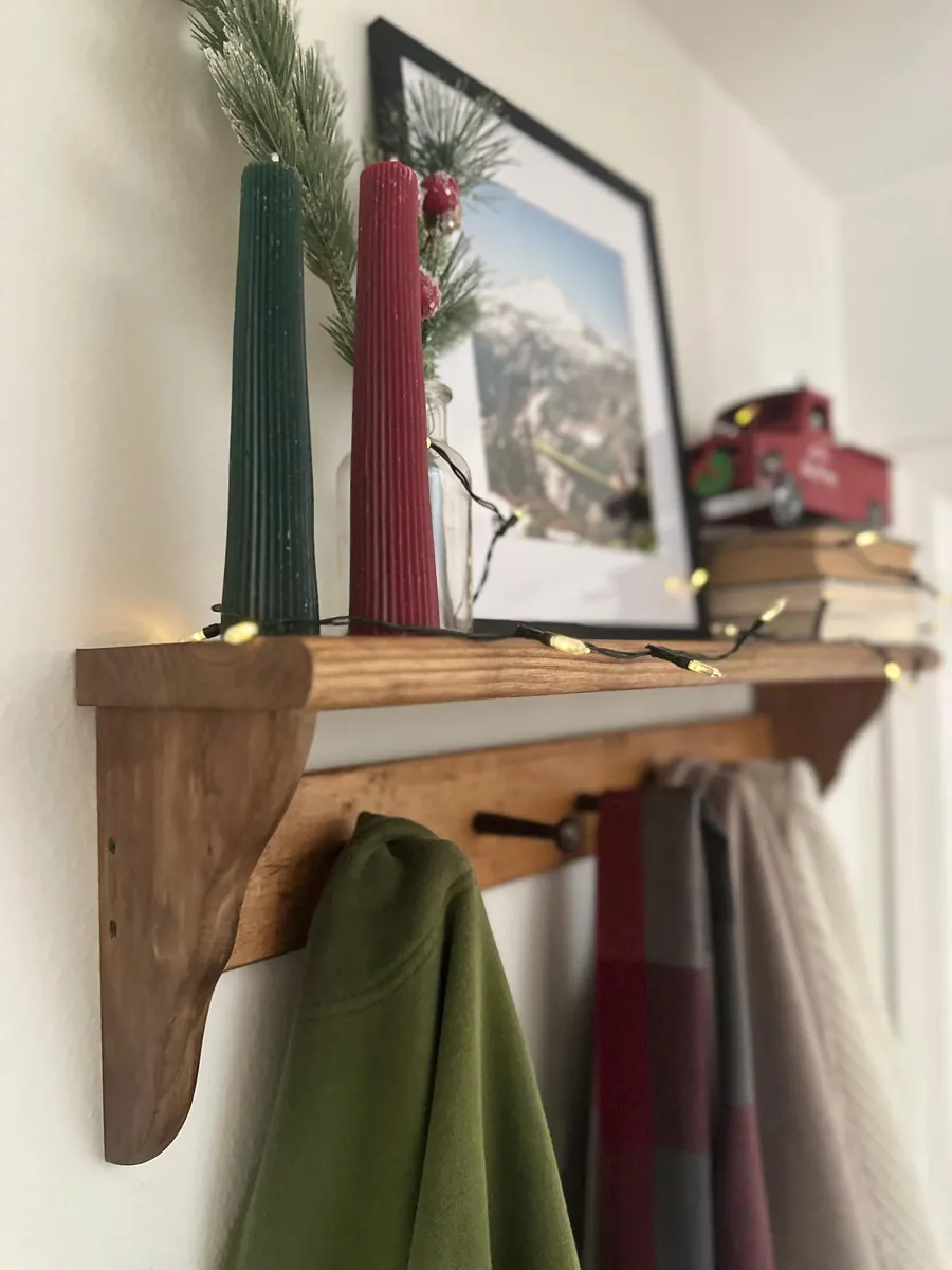 A peg shelf decorated for Christmas with candles, greenery, string lights and a small red truck, with coats and scarves on the pegs